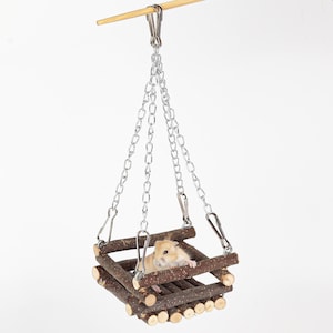 May include: A small, light brown hamster sits in a wooden swing suspended by silver chains. The swing is constructed from small, natural-looking wooden logs, creating a rustic appearance. The background is plain white.
