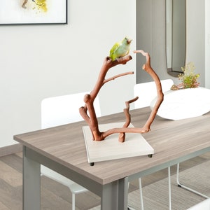 May include: A wooden bird perch with a natural branch design. A small, green and yellow bird is perched on one of the branches. The perch is mounted on a square, light-colored base.