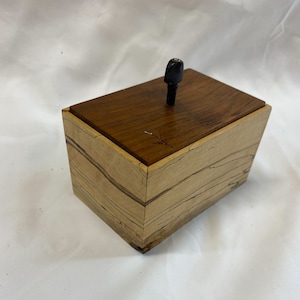 May include: A small wooden box with a light brown and dark brown wood grain pattern. The box has a hinged lid with a black knob on top.