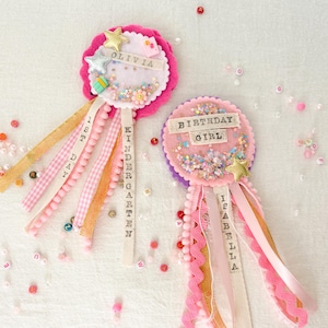 May include: Two circular felt badges with scalloped edges, featuring shaker centers filled with colorful beads and stars. One badge displays the name OLIVIA and 1ST DAY KINDERGARTEN, while the other displays BIRTHDAY GIRL and ISABELLA, with hanging ribbons.