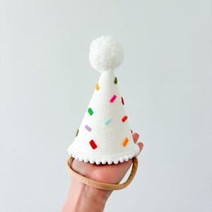 May include: A white party hat with colorful confetti-like accents and a white pom-pom on top. The hat has a scalloped edge and is attached to a tan headband. The hat is held in a hand against a white background.