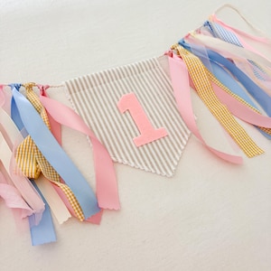 May include: A fabric pennant banner featuring a beige and white striped triangular flag with a large, soft pink felt number one centered on the front. The banner is decorated with various hanging ribbons in shades of pink, blue, and yellow gingham.