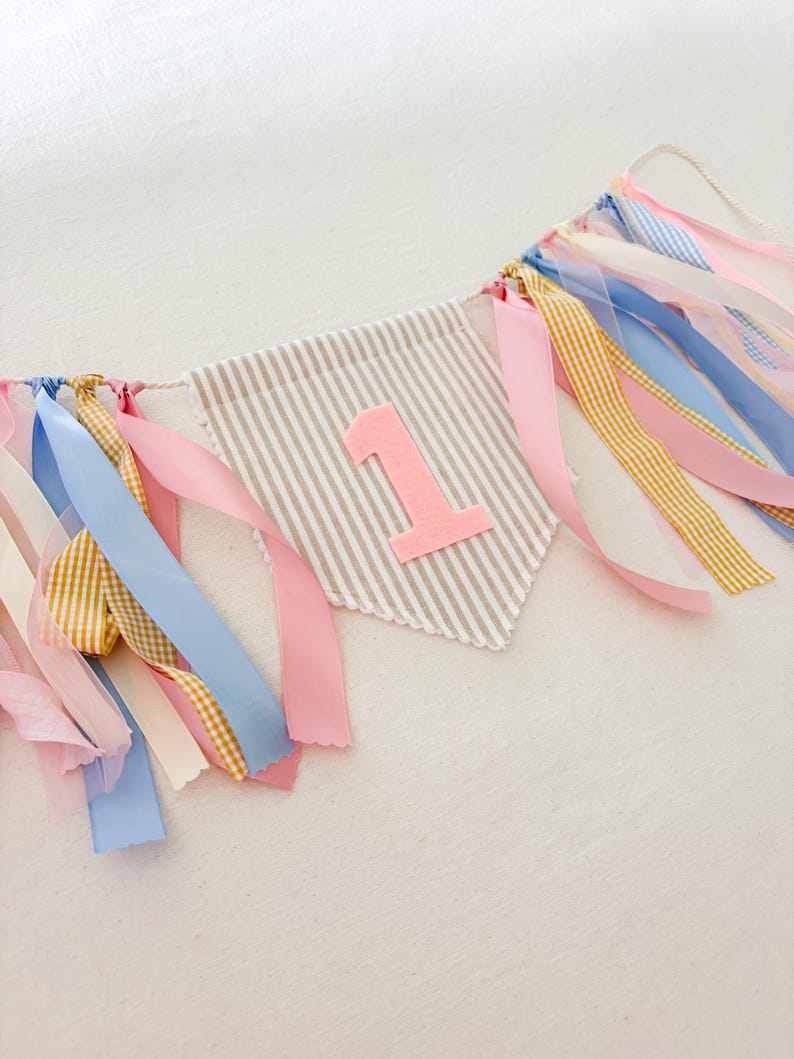 May include: A fabric pennant banner featuring a beige and white striped triangular flag with a large, soft pink felt number one centered on the front. The banner is decorated with various hanging ribbons in shades of pink, blue, and yellow gingham.