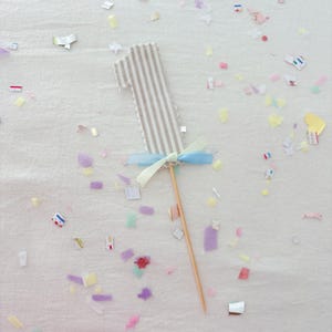 May include: A number one cake topper featuring a beige and white vertical striped fabric design with a scalloped edge. The topper is accented with a light blue and pale yellow ribbon bow tied at the base and is attached to a thin wooden stick.