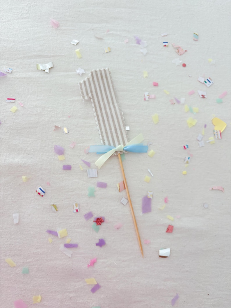 May include: A number one cake topper featuring a beige and white vertical striped fabric design with a scalloped edge. The topper is accented with a light blue and pale yellow ribbon bow tied at the base and is attached to a thin wooden stick.