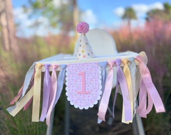 May include: A high chair banner featuring a scalloped center pennant with a pink number 1 and a colorful sprinkle pattern, flanked by hanging ribbons in pastel yellow, lavender, and blue gingham, paired with a matching party hat.