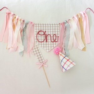 May include: A decorative banner featuring a beige and white gingham pennant with the word One in pink yarn, flanked by hanging fabric ribbons in pink, cream, and green. Includes a matching gingham number one cake topper and a plaid party hat.