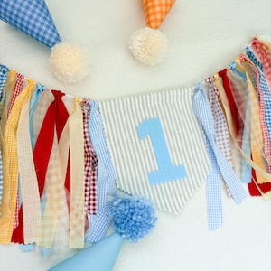 May include: A party banner featuring a central gray and white striped pennant with a light blue number 1, surrounded by hanging fabric ribbons in red, yellow, blue, and white gingham. Three cone-shaped party hats with pom-poms are included.