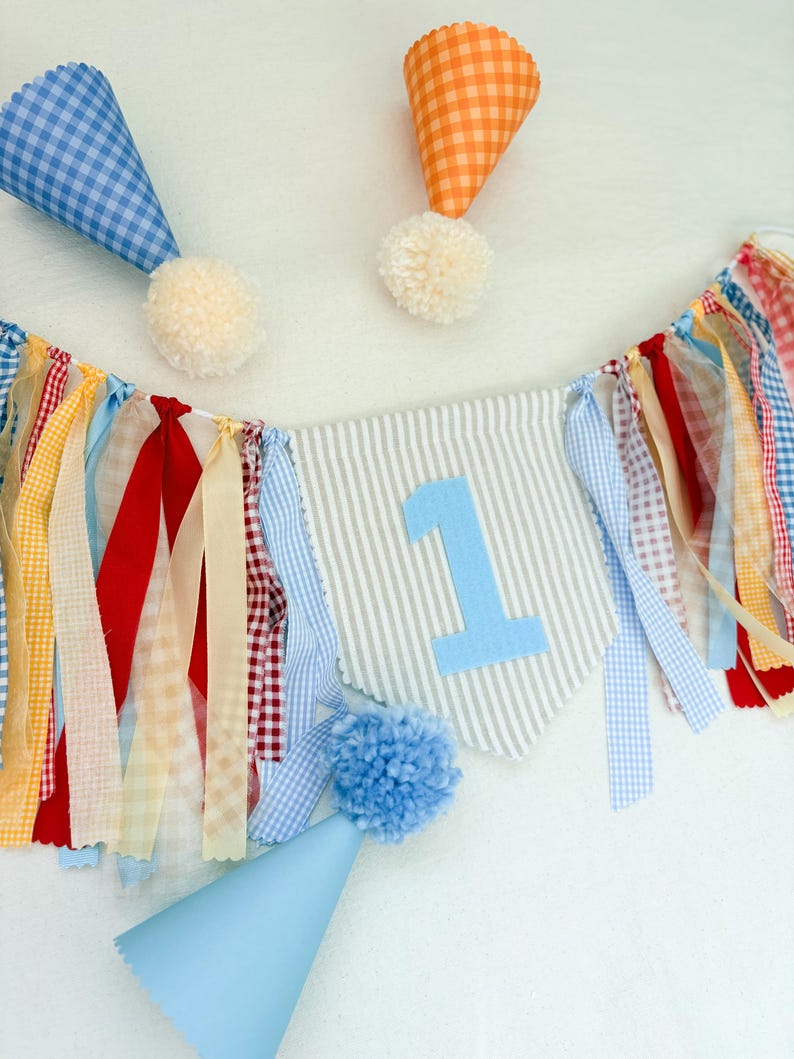 May include: A party banner featuring a central gray and white striped pennant with a light blue number 1, surrounded by hanging fabric ribbons in red, yellow, blue, and white gingham. Three cone-shaped party hats with pom-poms are included.