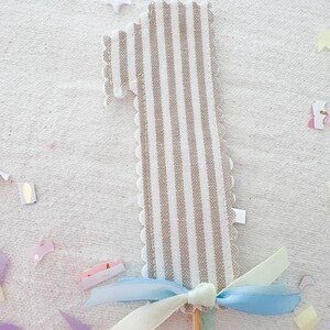 May include: A fabric number one cake topper featuring a beige and white vertical striped pattern with scalloped edges. The base of the number is decorated with a light blue and pale yellow ribbon tied in a bow, attached to a thin wooden stick.