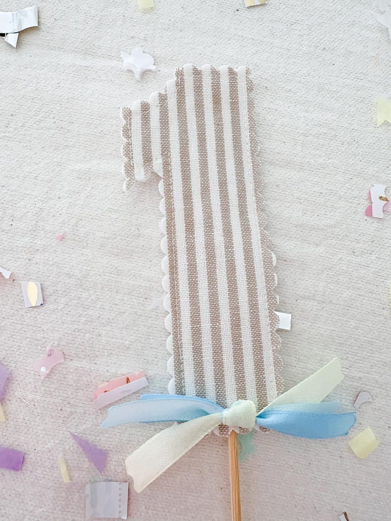 May include: A fabric number one cake topper featuring a beige and white vertical striped pattern with scalloped edges. The base of the number is decorated with a light blue and pale yellow ribbon tied in a bow, attached to a thin wooden stick.