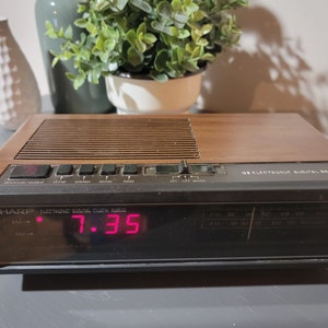 May include: A vintage brown wood-grain Sharp electronic digital clock radio with a red digital display showing the time 7:35. The clock radio has a variety of buttons including sleep, slow, fast, on, off, and auto.