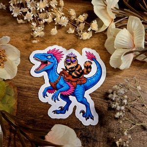Peut inclure: Un autocollant découpé représentant un dinosaure bleu vif avec des plumes roses et une selle marron. Un chat tigré brun et duveteux portant une petite couronne argentée et des lunettes de soleil violettes est assis sur la selle.