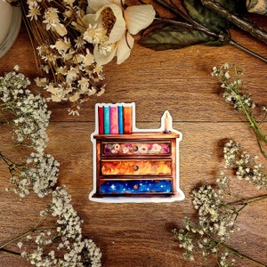 May include: A sticker of a wooden chest of drawers with three drawers. The drawers are decorated with flowers, flames, and a starry night sky. There are books on top of the chest of drawers and a candle on the right side.