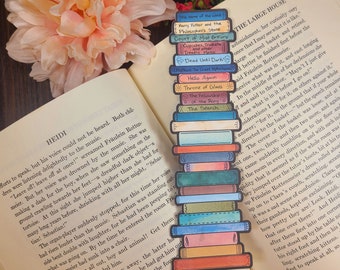 May include: A watercolor bookmark featuring a stack of colorful books with titles like "The Name of the Wind", "Harry Potter and the Philosopher's Stone", "Court of Mist & Fury", "Dead Until Darks", "Throne of Glass", "The Fellowship of the Ring", and "The Search".