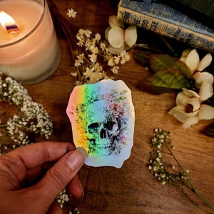 May include: A holographic sticker featuring a white skull with a floral design. The skull is surrounded by pink, yellow, and blue flowers. The sticker has a rainbow iridescent background.