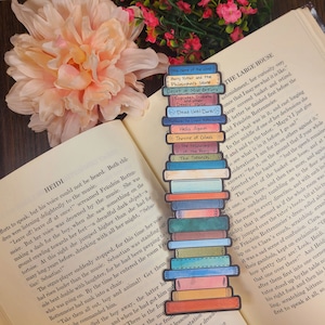May include: A watercolour bookmark featuring a stack of colourful books with titles such as "The Name of the Wind", "Harry Potter and the Philosopher's Stone", "Court of Mist & Fury", "Dead Until Darks", "Throne of Glass", "The Fellowship of the Ring", and "The Search".