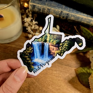 May include: A sticker of the state of West Virginia with a watercolor painting of a waterfall and forest inside the outline. The sticker has a white border and the words "West Virginia" are written in black cursive at the bottom.