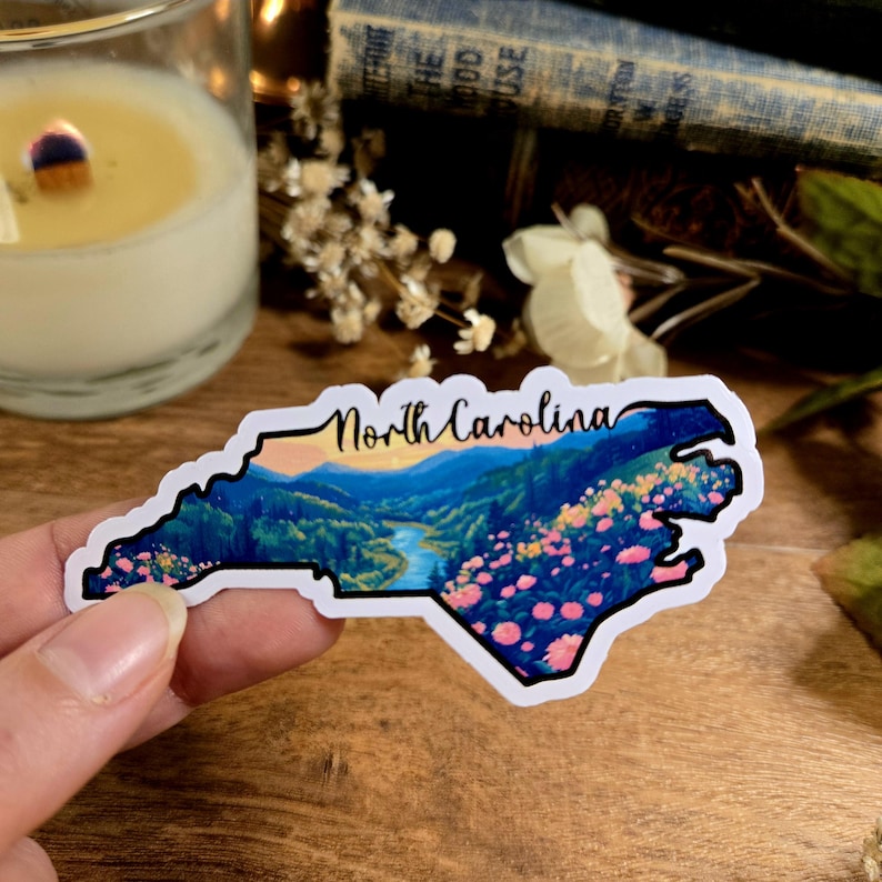 Puede incluir: Una pegatina con una ilustraci&oacute;n colorida del estado de Carolina del Norte con una cordillera y un r&iacute;o. La pegatina tiene un fondo blanco y las palabras "North Carolina" est&aacute;n impresas en negro en la parte superior.