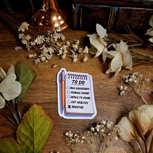 Op de afbeelding: Een sticker met een notitieblok met de woorden "TO DO" en een takenlijst: "Buy groceries", "Charge phone", "Reply to email", "Eat healthy" en "Breathe". Een oranje pen ligt naast het notitieblok.