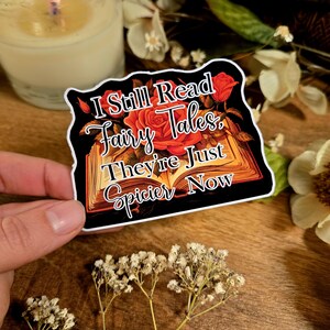 May include: A sticker with a black background and white border featuring an open book with red roses and the text "I Still Read Fairy Tales. They're Just Spicier Now."