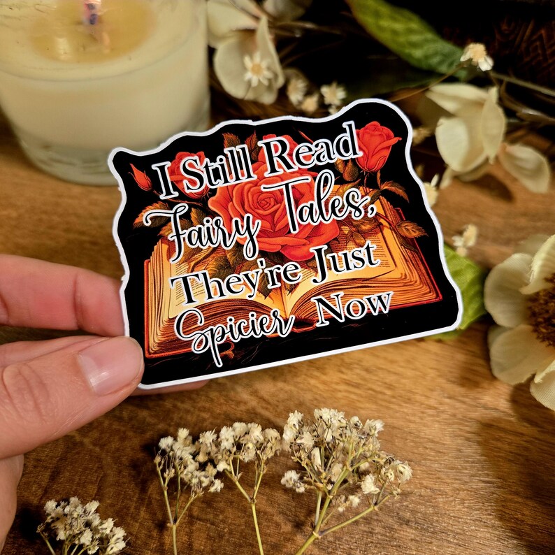 May include: A sticker with a black background and white border featuring an open book with red roses and the text "I Still Read Fairy Tales. They're Just Spicier Now."