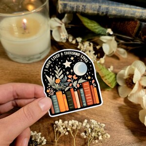 May include: A sticker with a black background and white border featuring a crescent moon, stars, and a stack of books. The text "I have lived a thousand lives" is written in a curved arch above the books.