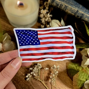 Puede incluir: Una pegatina de bandera americana roja, blanca y azul con efecto acuarela. La pegatina est&aacute; sobre una superficie de madera con flores y una vela.