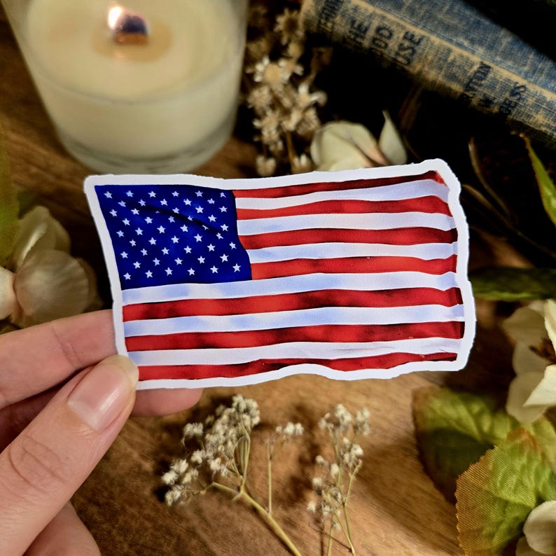 Puede incluir: Una pegatina de bandera americana roja, blanca y azul con efecto acuarela. La pegatina est&aacute; sobre una superficie de madera con flores y una vela.