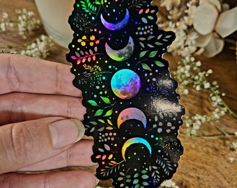 May include: A holographic sticker with a black background featuring a series of iridescent moons and leafy vines in rainbow colors.