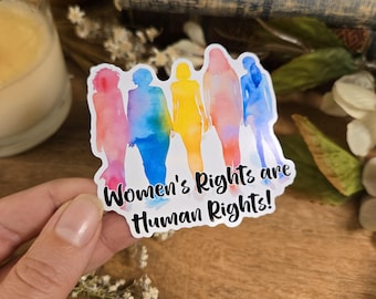 May include: A white sticker with a watercolor illustration of five figures in a rainbow color scheme. The text "Women's Rights are Human Rights!" is printed in black below the figures.