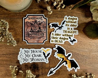 Peut inclure: Quatre autocollants sur le thème de la fantasy. Un autocollant représente une enseigne en bois avec les mots "Sorrengail Apothecary Dangerous Elixirs Brewed Daily". Un autre autocollant représente un dragon noir avec des accents jaunes et le texte "Un dragon sans son cavalier est une tragédie. Un cavalier sans son dragon est mort.". Un troisième autocollant représente un dragon noir avec des accents jaunes et le texte "Ma maison. Mon fauteuil. Ma femme.". Le dernier autocollant représente un dragon noir avec des accents jaunes.