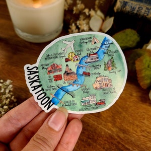 May include: A colourful illustrated map of Saskatoon, Saskatchewan, Canada. The map features landmarks, bridges, and points of interest. The map is on a white background with a blue river running through it.