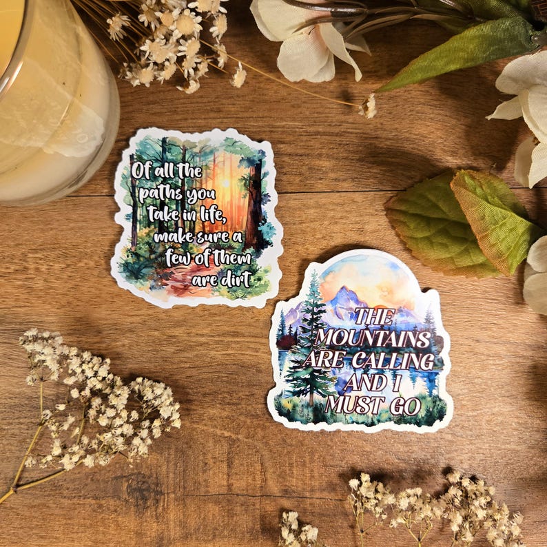 Puede incluir: Dos pegatinas de acuarela con citas inspiradoras. La primera pegatina presenta una escena de bosque con la cita "De todos los caminos que recorras en la vida, aseg&uacute;rate de que algunos de ellos sean de tierra". La segunda pegatina presenta una escena de monta&ntilde;a con la cita "Las monta&ntilde;as llaman y debo ir".