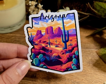 Puede incluir: Una pegatina con una colorida ilustración del paisaje de Arizona, incluyendo cactus, mesas y una puesta de sol. La palabra "Arizona" está escrita en cursiva en la parte superior. La pegatina está sujeta por una mano.
