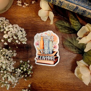 May include: A white sticker with a floral design and the text "I have no shelf control". The sticker features a stack of colorful books with a floral design.
