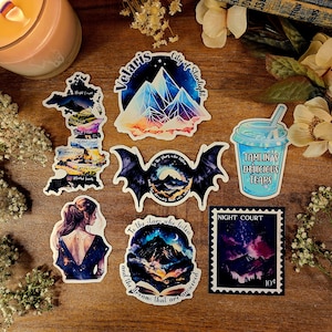 K&ouml;nnte beinhalten: Ein Set mit 7 bunten Aufklebern mit Illustrationen aus der Reihe A Court of Thorns and Roses. Die Aufkleber zeigen eine Karte von Prythian, eine Bergkette, eine Fledermaus, eine Tasse mit blauer Fl&uuml;ssigkeit, eine Bergkette mit einem Halbmond und eine Briefmarke mit einer Bergkette und einem Sternenhimmel. Der Text auf den Aufklebern lautet "Velaris City of Starlight", "Tamlin's Delicious Tears", "To the stars who listen and the dreams that are answered", und "Night Court 10&cent;".