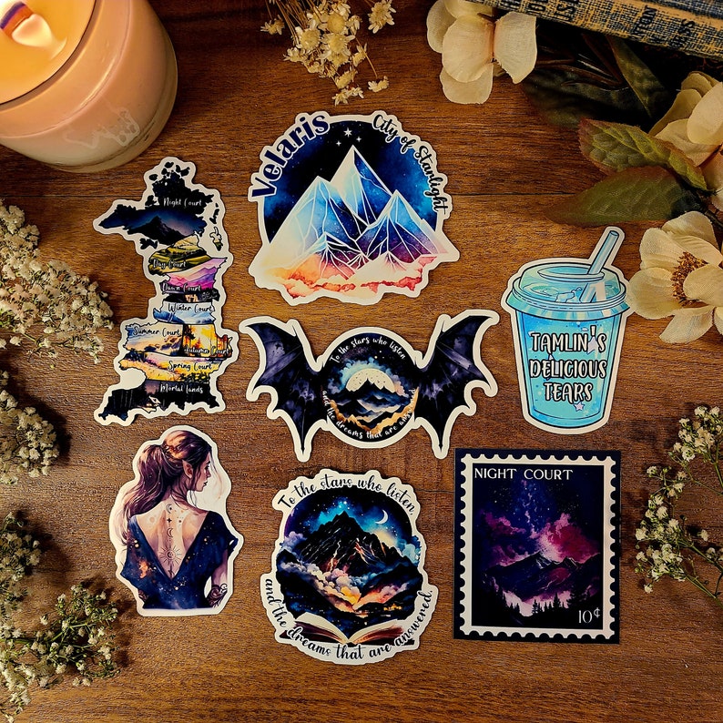 K&ouml;nnte beinhalten: Ein Set mit 7 bunten Aufklebern mit Illustrationen aus der Reihe A Court of Thorns and Roses. Die Aufkleber zeigen eine Karte von Prythian, eine Bergkette, eine Fledermaus, eine Tasse mit blauer Fl&uuml;ssigkeit, eine Bergkette mit einem Halbmond und eine Briefmarke mit einer Bergkette und einem Sternenhimmel. Der Text auf den Aufklebern lautet "Velaris City of Starlight", "Tamlin's Delicious Tears", "To the stars who listen and the dreams that are answered", und "Night Court 10&cent;".