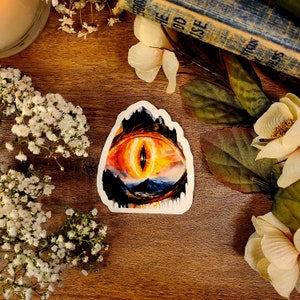 May include: A sticker featuring a large, fiery orange eye with a black pupil and a dark, rocky landscape surrounding it. The eye is set against a background of a mountain range and a body of water.
