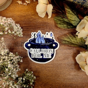 May include: A white sticker with a blue cauldron with a blue flame and the text "Cauldron Boil Me" on a wooden surface.