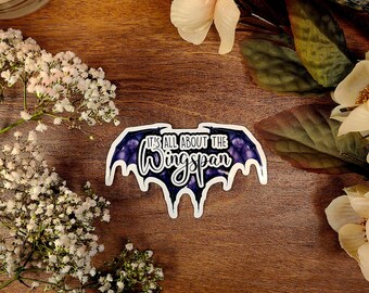 May include: A sticker with a purple bat design and the text "It's all about the Wingspan" on a wooden surface.