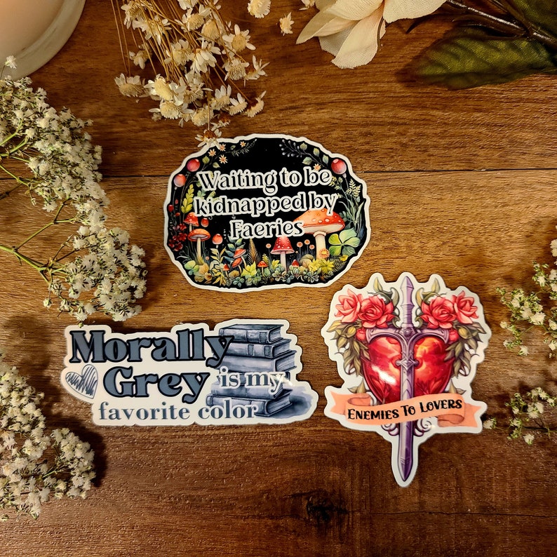May include: Three illustrated stickers with whimsical designs. The first sticker features a dark background with mushrooms and greenery and the text "Waiting to be kidnapped by Faeries". The second sticker features a stack of books with the text "Morally Grey is my favorite color". The third sticker features a red heart with a sword through it and the text "Enemies to Lovers".