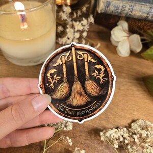 Könnte beinhalten: Ein runder Aufkleber mit braunem und goldenem Design. Der Aufkleber zeigt drei Besen mit goldenem Griff und braunen Borsten. Der Text "The Three Broomsticks" ist in goldener Schrift um den unteren Rand des Aufklebers geschrieben.