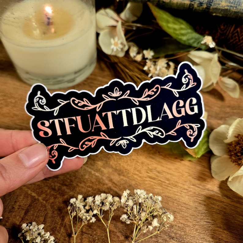 May include: A black and white sticker with a pink and white gradient design. The sticker features a decorative floral border and the text "STFUATTDLAGG".