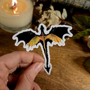 Peut inclure: Un autocollant de dragon noir et jaune avec les ailes déployées. Le dragon vole, la queue traînant derrière lui.