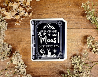 May include: A black and white sticker of a book cover with the text "Survivor of Maas Destruction". The book cover features a mountain range, a crown, a sword, a crescent moon, a deer, and a star.