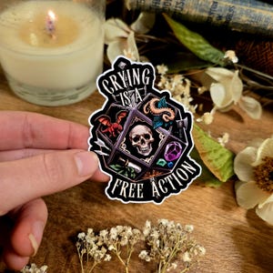 Puede incluir: Una pegatina negra con el texto "Crying is a Free Action". La pegatina presenta una calavera, un dragón rojo, una serpiente y otros elementos de fantasía. La pegatina está sujeta por una mano.