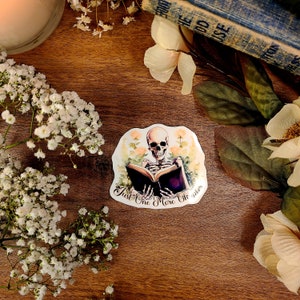 May include: A sticker featuring a skeleton reading a book with the text "Just One More Chapter". The skeleton is surrounded by flowers and the sticker has a white background.