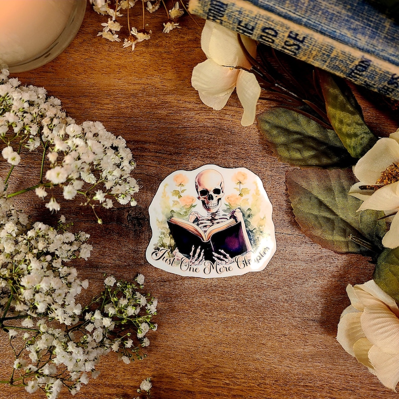 May include: A sticker featuring a skeleton reading a book with the text "Just One More Chapter". The skeleton is surrounded by flowers and the sticker has a white background.