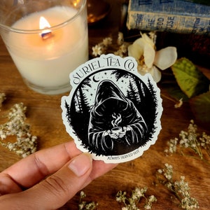 May include: A black and white sticker featuring a hooded figure holding a steaming cup of tea. The sticker reads "Suriel Tea Co. Always Served Hot" and includes a crescent moon and stars in the background.